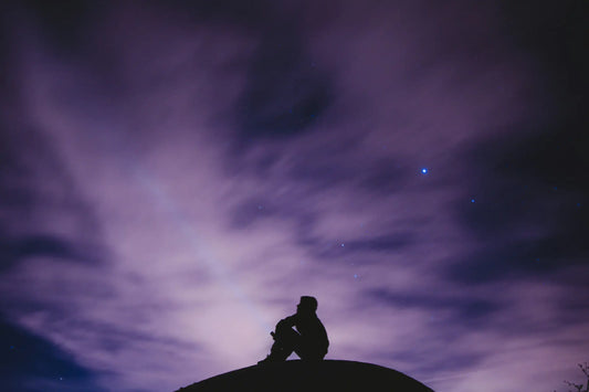 A silhouette of a person siting on a boulder with a dark purple cloudy sky in the background. The person's head is looking upward toward the sky.